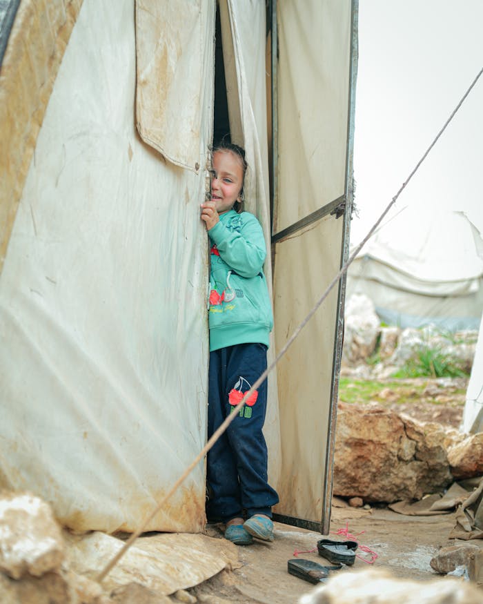 A young girl with blonde hair peeks out from a tent in a Syrian refugee camp, capturing innocence and resilience.
