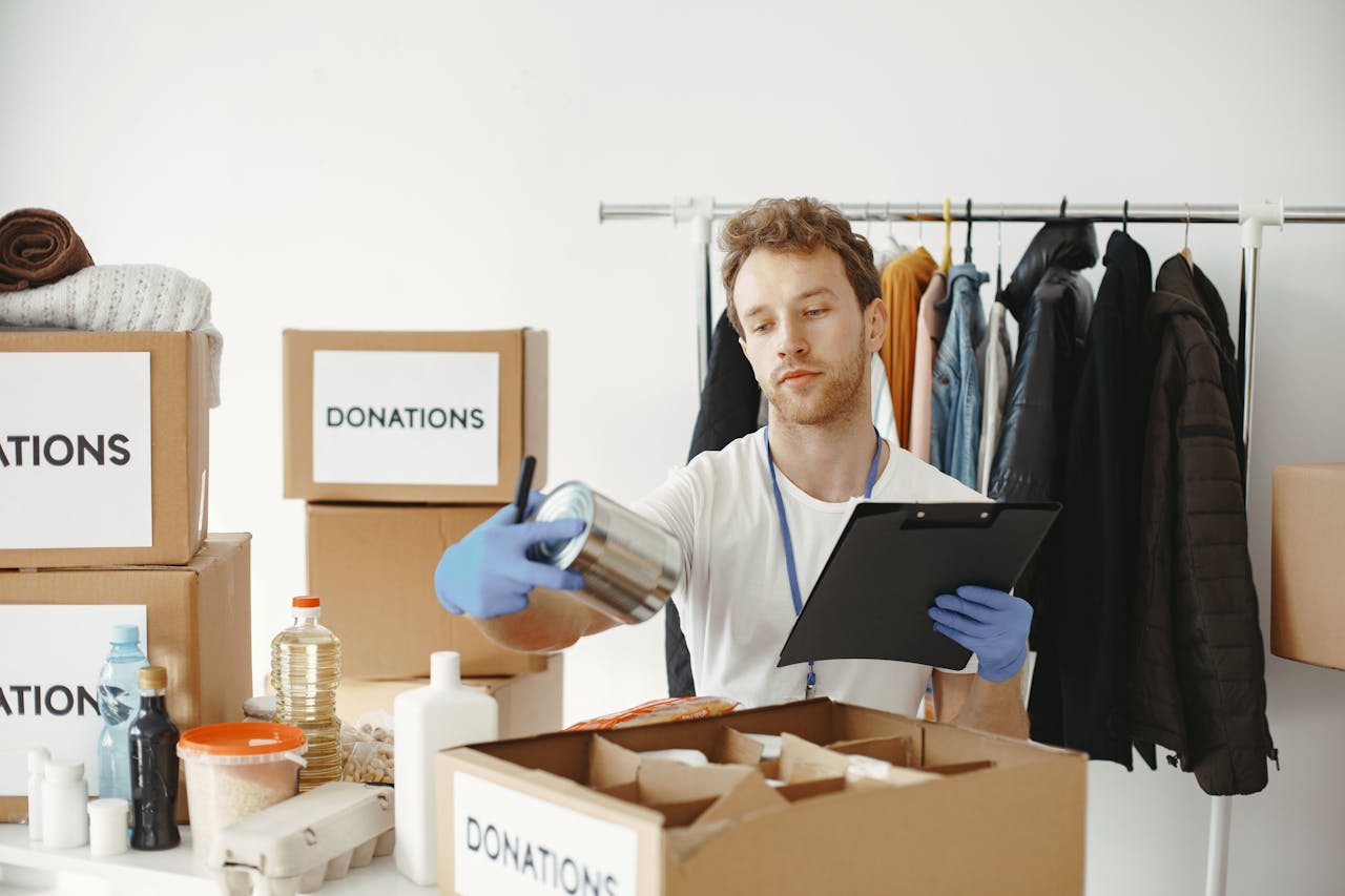 A volunteer organizes food and clothing donations in a community center, maintaining records on a clipboard.